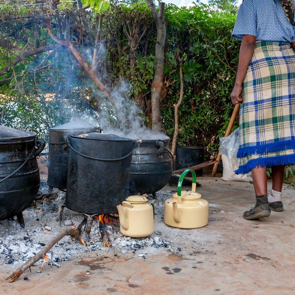 Photo depicts a woman cooking using four black cauldrons over an ashy and wood filled fire