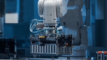 Photo depicts a Modern EV Factory. On Automated Production Line Robot Arms Transporting Automotive Battery Modules onto Conveyor Belt. Electric Car Battery Pack Manufacturing Process.